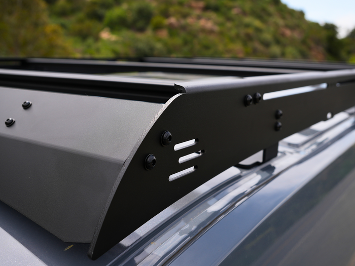 A modern high-roof van with a loaded flat platform roof rack carrying a spare wheel and cases, at golden hour