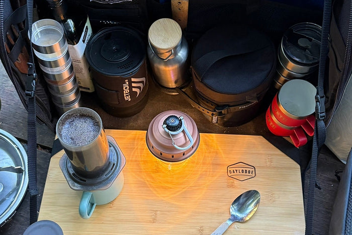 A fold-out camp kitchen case open on gravel beside a van at dawn with a steel pot steaming on the burner